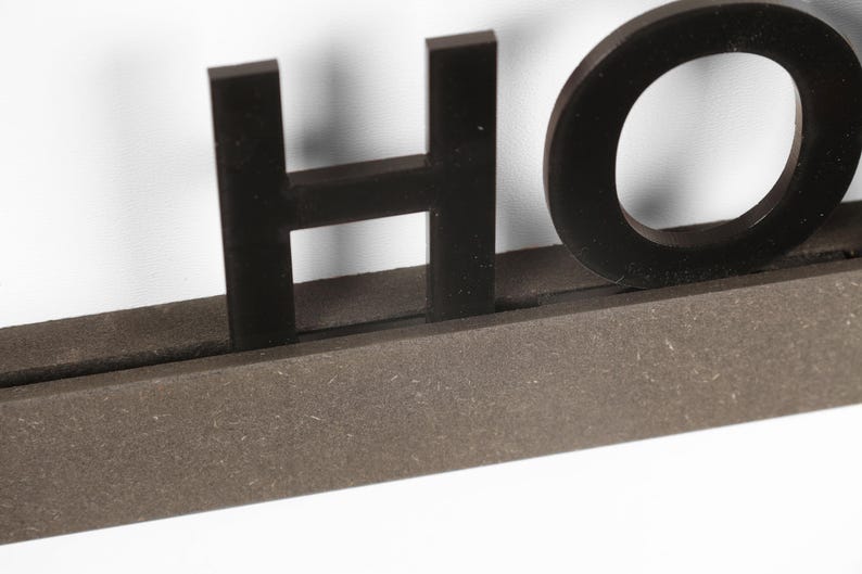 Puede incluir: Letras de madera negras que deletrean "HOME" montadas sobre una base de madera marr&oacute;n.