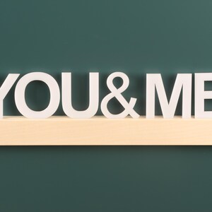 May include: White wooden letters spelling "YOU & ME" on a light brown wooden shelf against a dark green background.