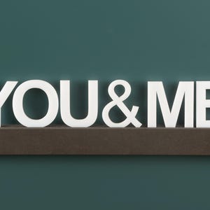 May include: White wooden letters spelling "YOU & ME" are displayed on a dark brown shelf against a teal green background.