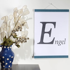 May include: A white poster with a black letter E and the word "engel" printed on it. The poster is hanging on a white wall with a blue frame. A vase with white flowers is in the foreground.