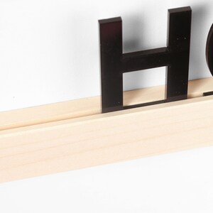 Puede incluir: Letras negras de acr&iacute;lico que deletrean "HOME" se muestran en un estante de madera natural.