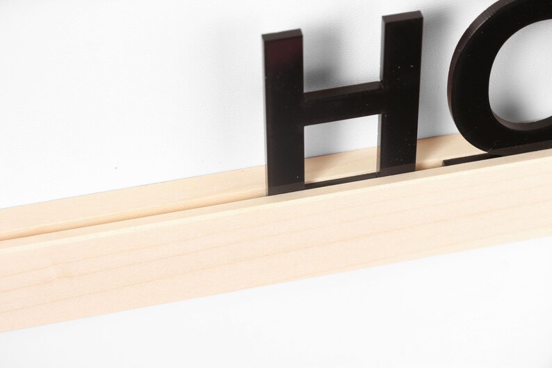 Puede incluir: Letras negras de acr&iacute;lico que deletrean "HOME" se muestran en un estante de madera natural.