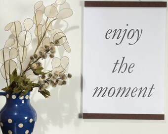 May include: A white framed print with the text "enjoy the moment" hanging on a white wall. A vase with a blue and white polka dot pattern holds dried flowers.
