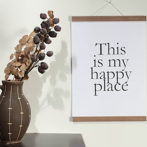 May include: A white framed print with the text "This is my happy place" hanging on a white wall. A brown vase with a dried floral arrangement is in the foreground.