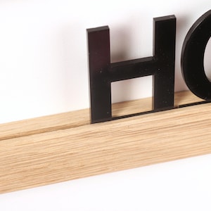 Puede incluir: Letras negras de acr&iacute;lico que deletrean "HOME" montadas en un estante de madera marr&oacute;n claro.