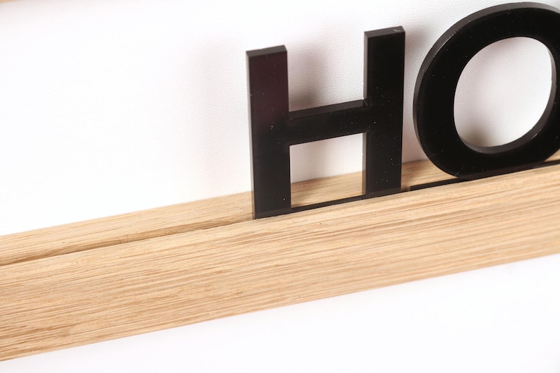 Puede incluir: Letras negras de acr&iacute;lico que deletrean "HOME" montadas en un estante de madera marr&oacute;n claro.