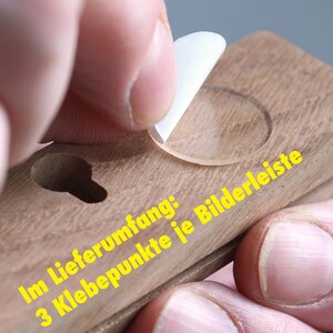 Puede incluir: Primer plano de un marco de fotos de madera con un c&iacute;rculo de pl&aacute;stico transparente en la superficie. El texto "Im Lieferumfang: 3 Klebepunkte je Bilderleiste" es visible en el marco.