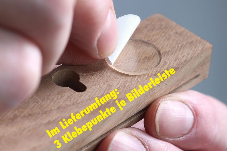 Puede incluir: Primer plano de un marco de fotos de madera con un c&iacute;rculo de pl&aacute;stico transparente en la superficie. El texto "Im Lieferumfang: 3 Klebepunkte je Bilderleiste" es visible en el marco.