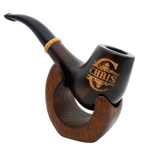 Könnte beinhalten: Eine dunkelbraune Holzpfeife mit schwarzem Stiel und einem Holzständer. Die Pfeife hat ein goldenes, graviertes Design mit dem Namen "CHRIS" darauf.