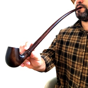 Könnte beinhalten: Eine dunkelbraune, gebogene Tabakpfeife mit einem abgerundeten Kopf und einem langen, schlanken Stiel. Die Pfeife wird in der Hand einer Person gehalten. Die Pfeife ist aus Holz und hat eine polierte Oberfläche. Die Person trägt ein kariertes Hemd.