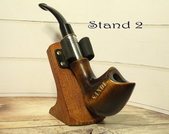 May include: A brown tobacco pipe with a curved stem, displayed on a wooden stand. The pipe is engraved with the word "SANDE". The stand is made of wood and has the text "Stand 2" in the background.