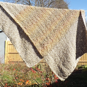 May include: A beige and brown knitted throw blanket with a gradient pattern. The blanket is hanging on a clothesline outdoors.