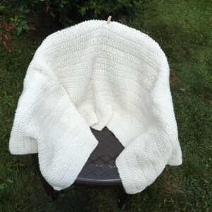 May include: A white crocheted blanket draped over a brown plastic chair. The blanket is made with a simple stitch pattern and has a soft, fluffy texture.