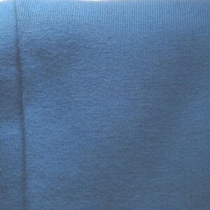 May include: Close-up of a textured, blue fabric. The fabric appears to be a knit or woven material, with visible texture and slight variations in color. The image is well-lit, highlighting the details of the textile.