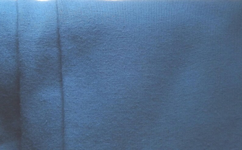 May include: Close-up of a textured, blue fabric. The fabric appears to be a knit or woven material, with visible texture and slight variations in color. The image is well-lit, highlighting the details of the textile.