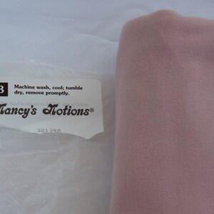 Puede incluir: Una etiqueta blanca con texto negro que dice "Machine wash, cool; tumble dry, remove promptly." y "Nancy's Notions&reg;" con el n&uacute;mero 321390 debajo. La etiqueta est&aacute; en una bolsa de pl&aacute;stico blanca. Un trozo de tela rosa est&aacute; a la derecha de la bolsa.