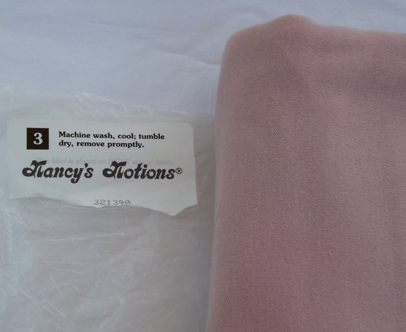 Puede incluir: Una etiqueta blanca con texto negro que dice "Machine wash, cool; tumble dry, remove promptly." y "Nancy's Notions&reg;" con el n&uacute;mero 321390 debajo. La etiqueta est&aacute; en una bolsa de pl&aacute;stico blanca. Un trozo de tela rosa est&aacute; a la derecha de la bolsa.
