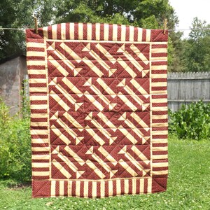 May include: A quilted throw blanket with a brown and cream geometric pattern. The blanket has a striped border with alternating brown and cream stripes.