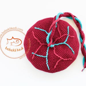 May include: A round, dark red crocheted pouch featuring a light blue stitched pattern on the surface and a braided handle made of dark red and light blue yarn. The text "bebeklikedi.com" and a cat logo are printed in orange on the left side.