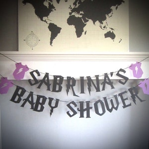 May include: Black and purple baby shower banner with the text "Sabrina's Baby Shower" in a gothic font. The banner is decorated with lightning bolt designs and purple baby onesies.