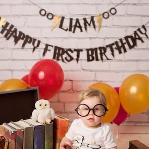 Puede incluir: Un bebé con gafas redondas negras y una camiseta blanca con un rayo y el texto "The Boy Who Lived" se sienta frente a una pila de libros. Un peluche de búho blanco se sienta encima de los libros. Una pancarta con el texto "Happy First Birthday Liam" cuelga sobre el bebé. La pancarta está decorada con rayos dorados y círculos. El fondo es una pared de ladrillo blanco con globos rojos, amarillos y rosas.