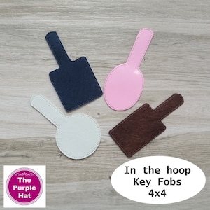 May include: Four key fobs in navy blue, light pink, white, and dark brown with textured surfaces. The navy blue and brown fobs are rectangular with tabs, while the pink and white fobs are circular with tabs. Text reads: In the hoop Key Fobs 4x4.