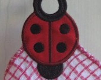 May include: A hanging kitchen towel featuring an embroidered ladybird applique at the top. The ladybird has a bright red body with black spots and a black head. The towel fabric is white with a red checkered pattern, and the entire piece is suspended from a black hook.