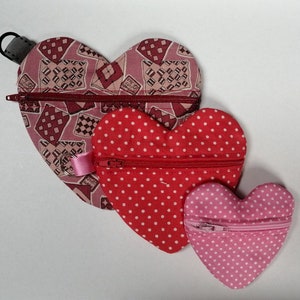 Può includere: Tre pochette con cerniera a forma di cuore di diverse dimensioni. La pochette più grande è rosa con un motivo a scacchi, la pochette di mezzo è rossa con pois bianchi e la pochette più piccola è rosa con pois bianchi.