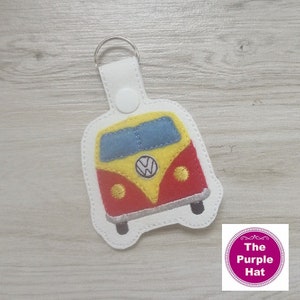 May include: A white felt keychain featuring an embroidered Volkswagen bus in red, yellow and blue. The keychain has a silver metal ring.