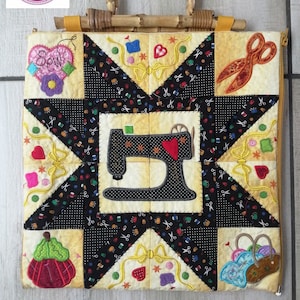 May include: A quilted sewing-themed tote bag with a bamboo handle. The bag features a patchwork design with a sewing machine, scissors, and other sewing notions. The color palette includes yellow, black, and red. The text "The Purple Hat" is in a circle in the upper left corner.