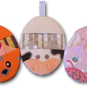 May include: Three oval-shaped fabric ornaments with hanging loops. The left is orange with a tiger face, the center is cream and brown with a cow face, and the right is pink with a pig face. Each features quilted patterns and stitched details.