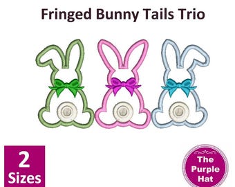 May include: Three embroidered bunny tail designs with fringed edges. Each bunny tail is a different color: green, pink, and blue. The bunny tails have a white center with a button-like detail. The designs are available in two sizes. The text "Fringed Bunny Tails Trio" and "The Purple Hat" are also included in the image.