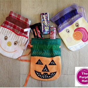 Op de afbeelding: Drie kleine stoffen zakjes met geborduurde gezichten. De zakjes zijn gevuld met snoep en zijn versierd met een geruit patroon, stippen en een pompoenontwerp. De zakjes zijn gelabeld met "The Purple Hat".