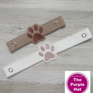 以下が含まれることがあります： ペットをテーマにした2つのリストバンド。1つは茶色い足跡が付いたタン色、もう1つはピンクの足跡が付いた白色です。各バンドには2つのシルバーのアイレットが付いています。The Purple Hatのロゴが右下にあります。