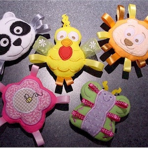 May include: A collection of five colorful, plush baby toys. The toys include a panda, a chick, a lion, a flower with a bee, and a butterfly. Each toy has ribbons in various colors.