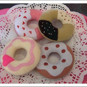 May include: Four colorful felt donuts on a pink plate with a white lace doily. The donuts have various designs, including hearts, sprinkles, and a heart-shaped accent. The donuts are in shades of pink, white, brown, and tan.