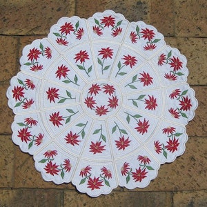 May include: Circular white poinsettia-themed placemat with a scalloped edge. The placemat features a repeating pattern of red poinsettia flowers with green leaves, arranged in a radial design. The design is embroidered on a white fabric.