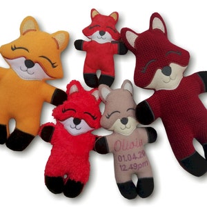 May include: Five plush fox toys in various shades of red, orange, and tan. Each fox has a smiling face with closed eyes, black paws, and a white muzzle. One fox has embroidered text: "Olivia 01.04.20 12.49pm".