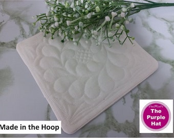 Könnte beinhalten: Ein quadratischer Untersetzer aus cremefarbenem Stoff mit aufwendigen, maschinell gesteppten Blumenmustern. Der Untersetzer zeigt den Text Made in the Hoop und ein kreisförmiges Logo mit dem Text The Purple Hat. Ein Zweig mit kleinen weißen Blüten liegt darauf.