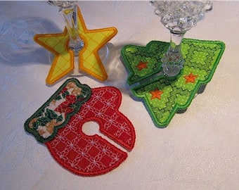 May include: Three embroidered fabric wine glass markers featuring festive shapes: a yellow star, a green Christmas tree, and a red ornament with a floral top section. Each marker has a slit to slide onto the stem of a glass for identification.