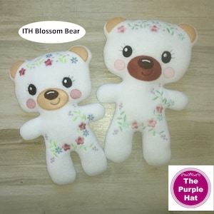 Puede incluir: Dos osos de peluche blancos con detalles florales bordados y narices marrones. Los osos están de pie sobre una superficie de madera. El texto "ITH Blossom Bear" es visible en la parte superior de la imagen.