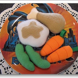 May include: A plate featuring a selection of felt food items, including a chicken leg, a carrot, green beans, and mashed potato. The plate has a cartoon image of Batman on it.