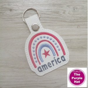 May include: A white vinyl keychain featuring an embroidered rainbow design with pink and light blue arches, a red star, and small blue star accents. The word america is embroidered in grey thread at the bottom. It includes a silver metal snap and ring.
