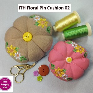 May include: Two pincushions, one brown and one pink, with floral embroidery. Both pincushions have a yellow button in the center. The image also includes two spools of embroidery floss, one yellow and one green, and a pair of gold scissors.