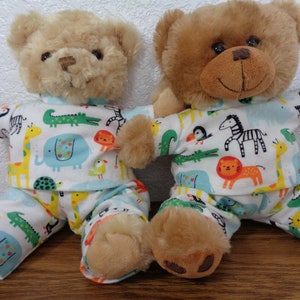 Pode incluir: Dois ursos de pelúcia vestindo pijamas combinando. Os pijamas são brancos com uma estampa colorida de animais de zoológico, incluindo girafas, elefantes e zebras. Os ursos são marrons claros e bege.