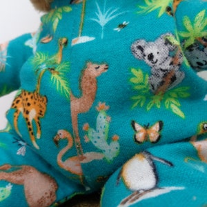 May include: A plush teddy bear wearing turquoise pajamas with a colorful print of animals and plants. The pattern includes giraffes, camels, flamingos, koalas, butterflies, and cacti. The bear's fur is light brown.