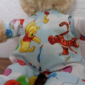Pode incluir: Um brinquedo de pelúcia vestindo um pijama azul claro com uma estampa colorida de Ursinho Pooh e Tigrão. O pijama apresenta ilustrações dos personagens, balões e pipas. O brinquedo tem pelo castanho e está posicionado com os braços estendidos.