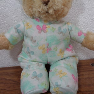 Pode incluir: Um urso de pelúcia vestindo um pijama azul claro com estampa de borboletas amarelas, rosas e cinzas. O urso tem pelo castanho e está sentado em uma superfície de madeira.