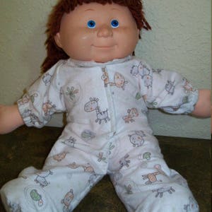 Puede incluir: Una muñeca de bebé vintage con cabello castaño y ojos azules, vestida con un mono blanco. El mono tiene un estampado de varios animales de dibujos animados en naranja, verde y gris. La muñeca está sentada con las piernas extendidas.