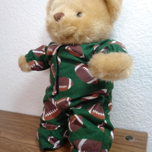 Pode incluir: Um urso de pelúcia bege vestindo um pijama verde com bolas de futebol americano marrons e brancas. O urso tem pelo castanho claro e um nariz castanho. O pijama tem um zíper na frente.